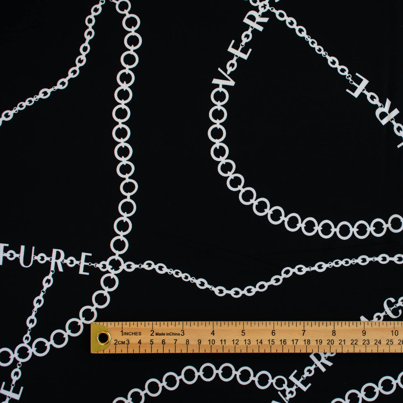 Italian High-End Designer Couture Knit in Black designer deadstock fabric by the yard. Classic \/ers@ce ITY jersey knit featuring white chains with the designer name and 'Couture' wording against a black background.  Image of fabric design scale with ruler.