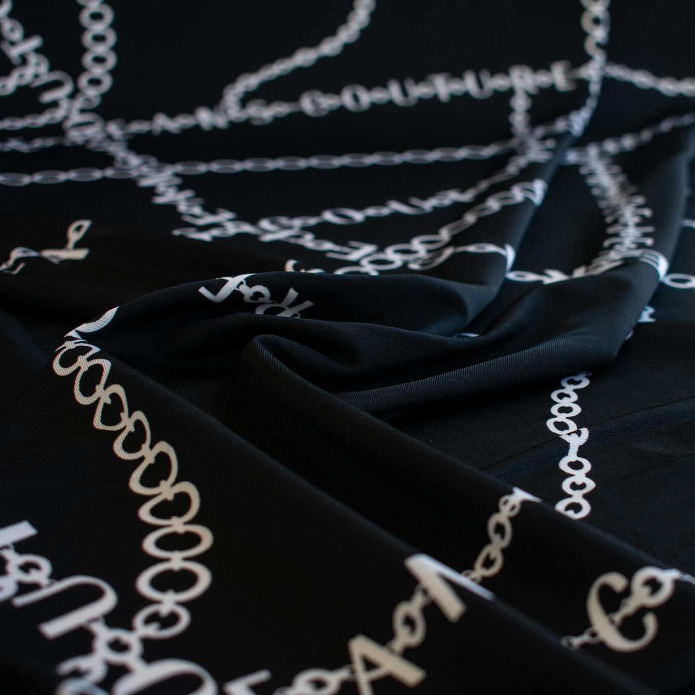 Italian High-End Designer Couture Knit in Black designer deadstock fabric by the yard. Classic \/ers@ce ITY jersey knit featuring white chains with the designer name and 'Couture' wording against a black background.
Close up image.