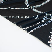 Italian High-End Designer Couture Knit in Black designer deadstock fabric by the yard. Classic \/ers@ce ITY jersey knit featuring white chains with the designer name and 'Couture' wording against a black background. Image of selvedge.