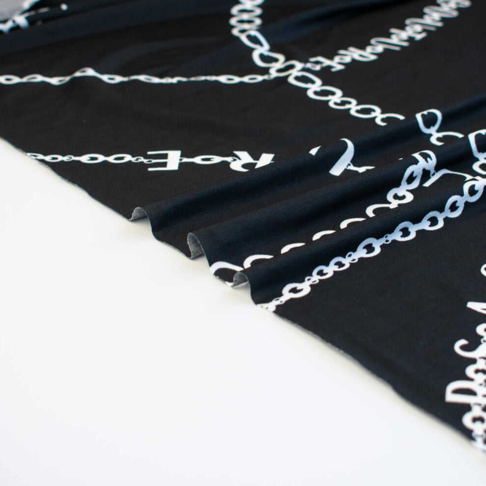 Italian High-End Designer Couture Knit in Black designer deadstock fabric by the yard. Classic \/ers@ce ITY jersey knit featuring white chains with the designer name and 'Couture' wording against a black background. Image of selvedge.