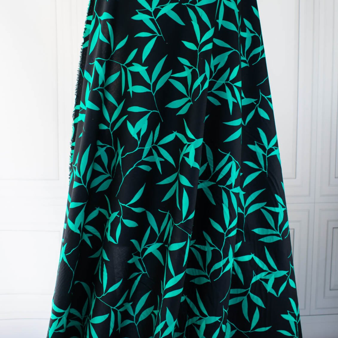 Carolina' cotton poplin designer fabric by the yard. This Italian poplin is light and billowy, perfect for sewing up tops, dresses and skirts. Made with stretch for ultimate comfort, this fabric is perfect if you want to add a touch of the exotic with its turquoise bamboo leaf print set against a black background. Image of fabric drape.
