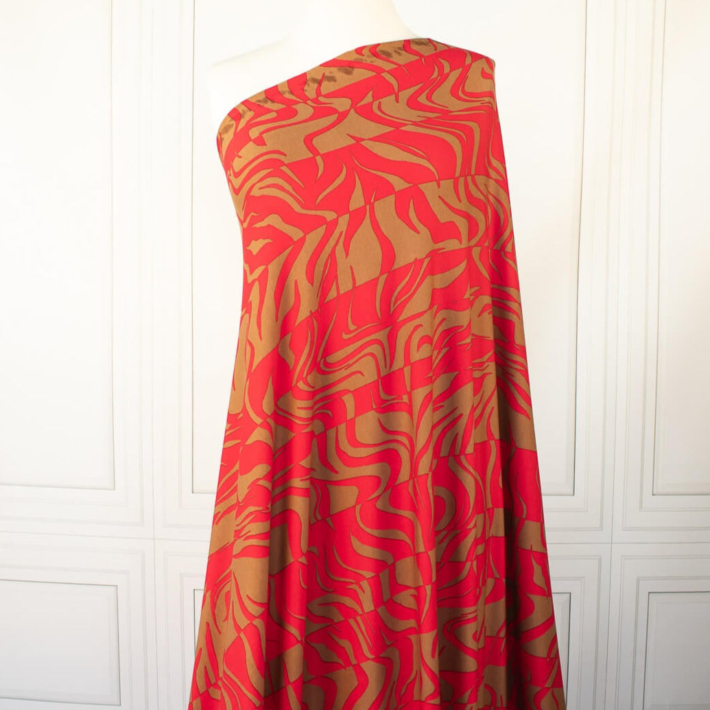 Italian Single Viscose Jersey Red and Caramel Modern Print fabric by the yard. A gorgeous vibrant knit that is sure to capture compliments. We love this combination of red and caramel brown tones in this modern jersey knit fabric. The printed design has a bit of animal print edge in a stripe like pattern. Longer image of fabric draped on dressform.