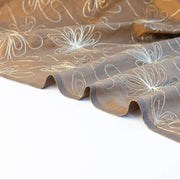 100% silk shantung fabric by the yard featuring a metallic embroidered floral design in a modern and playful hand-drawn style. In a rich hazelnut brown hue, this fabric offers a crisp and light textured hand, with a soft sheen for added elegance. Image of selvedge.