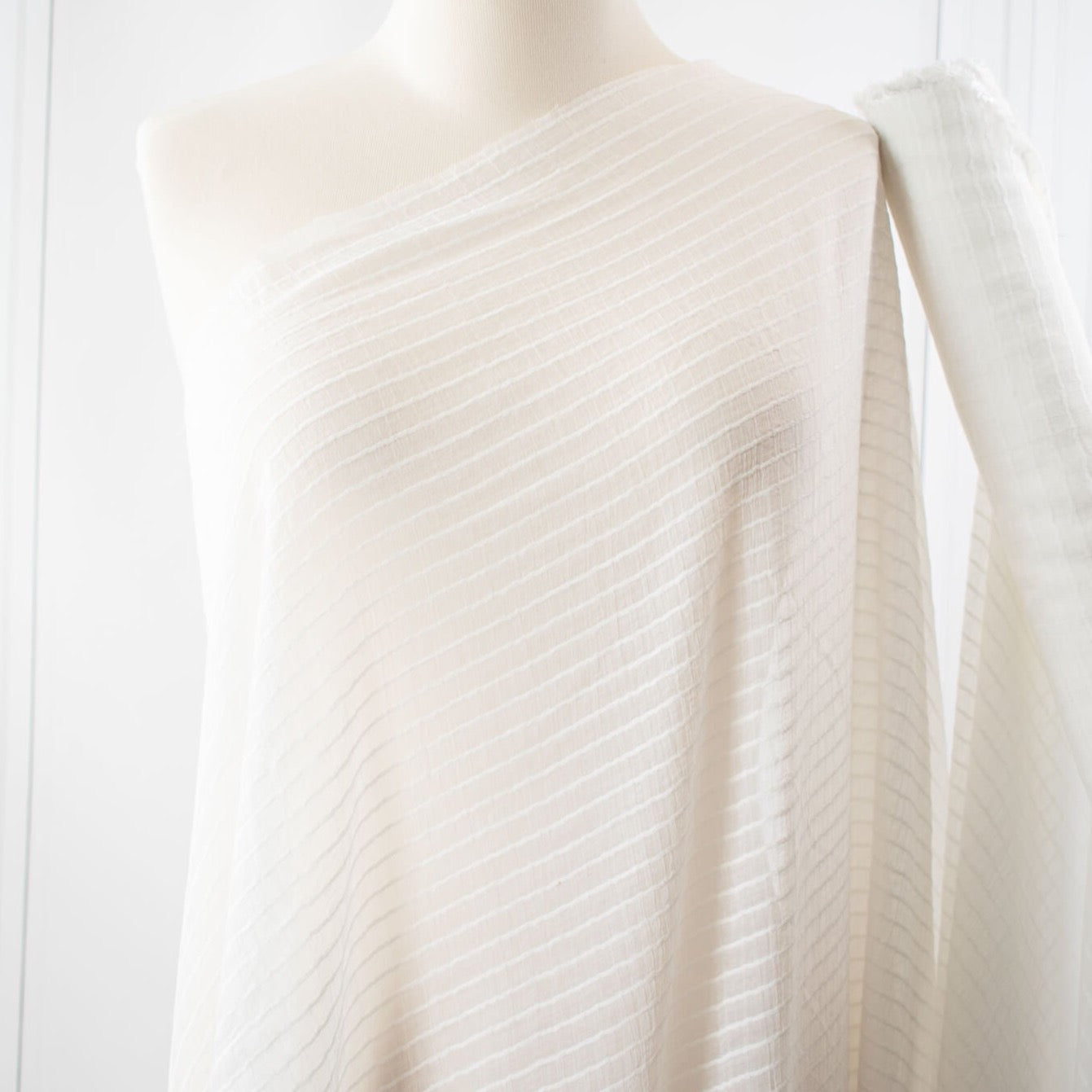 You will be dreaming of summer in this soft, sheer designer woven fabric.  This feels so good against the skin, a must have for summer dresses and tops. Gorgeous sewn up in a loose fitting garment or take advantage of the stripe and go for a bias cut!  Fabric has a white jacquard horizontal stripe against a white sheer background.  Image of fabric draped on mannequin.