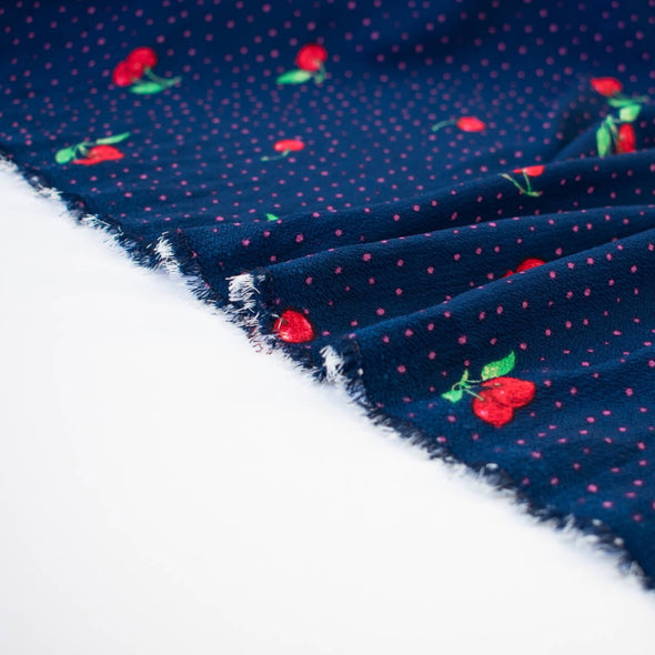Have some fun with our navy and pink polka dot designer deadstock fabric by the yard adorned with vibrant red cherries. Made from high-quality polyester crepe, this fabric has a textured hand, lovely drape, and a vintage vibe. Image of fabric selvedge.