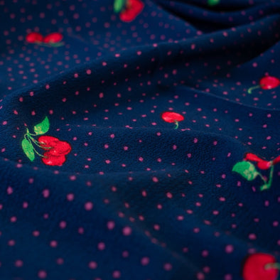 Have some fun with our navy and pink polka dot designer deadstock fabric by the yard adorned with vibrant red cherries. Made from high-quality polyester crepe, this fabric has a textured hand, lovely drape, and a vintage vibe. Close up image.