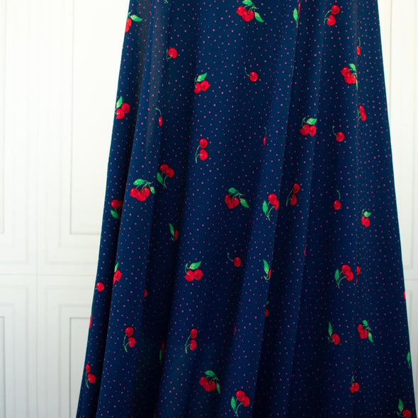 Have some fun with our navy and pink polka dot designer deadstock fabric by the yard adorned with vibrant red cherries. Made from high-quality polyester crepe, this fabric has a textured hand, lovely drape, and a vintage vibe. Image of fabric drape.