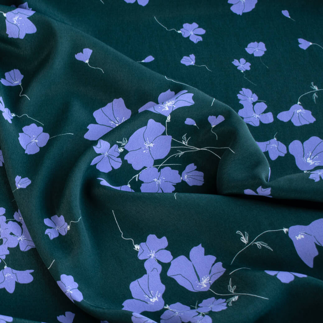 Close up of fabric design. Italian Floral Viscose Crepe designer deadstock fabric by the yard. Flowers of French Blue wisp across a deep green background that evokes a breezy mood of pleasant days.