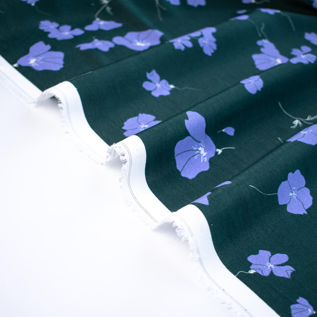 Image of fabric selvedge. Italian Floral Viscose Crepe designer deadstock fabric by the yard. Flowers of French Blue wisp across a deep green background that evokes a breezy mood of pleasant days.