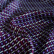 Close up image of Italian Viscose Satin Modern Geometric Print designer deadstock fabric by the yard. From the Italian mills, enjoy a sophisticated geometric print of small squares in shades of purple highlighted by an ivory background. A cool smooth hand and satiny sheen is perfect for creating a top or dress that can take you from day to evening.