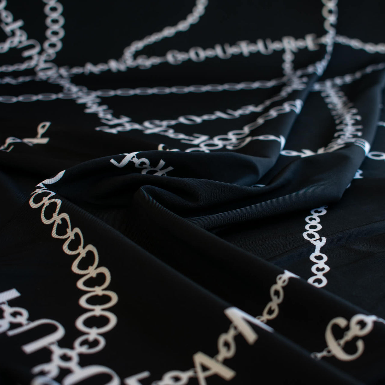 Italian High-End Designer Couture Knit in Black designer deadstock fabric by the yard. Classic \/ers@ce ITY&nbsp;jersey knit featuring white chains with the designer name and 'Couture' wording against a black background.
Close up image.