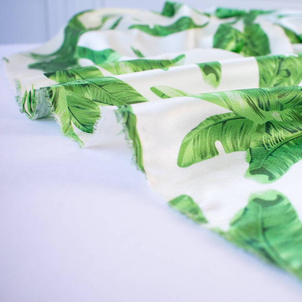 Designer Silk/Lycra Charmeuse Palm Print fabric by the yard! This fun and fashion forward deadstock fabric is perfect for creating unique and trending pieces. The green palm print set against a soft white background and the lustrous finish of this fabric will elevate any outfit.  Image of fabric selvedge.