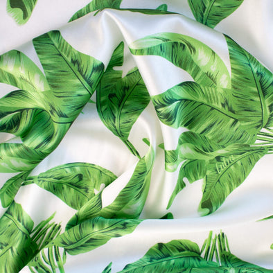 Designer Silk/Lycra Charmeuse Palm Print fabric by the yard! This fun and fashion forward deadstock fabric is perfect for creating unique and trending pieces.  The green palm print set against a soft white background and the lustrous finish of this fabric will elevate any outfit. Close up image.