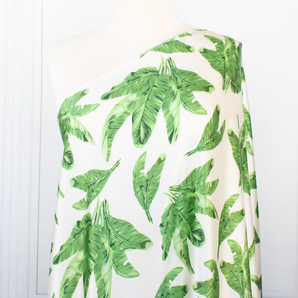 Designer Silk/Lycra Charmeuse Palm Print fabric by the yard! This fun and fashion forward deadstock fabric is perfect for creating unique and trending pieces. The green palm print set against a soft white background and the lustrous finish of this fabric will elevate any outfit.  Image of fabric scale on dressform.