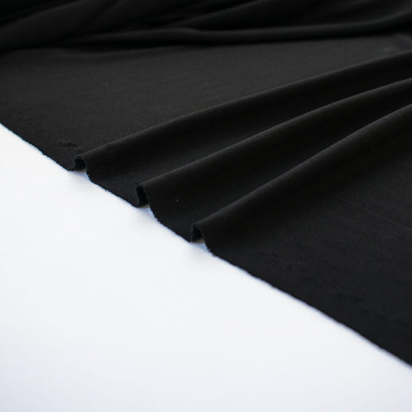 High-end Designer Black Matte jersey knit deadstock fabric by the yard, a timeless essential for every wardrobe. Achieve a designer-level look and feel, with a sophisticated black matte finish. Image of fabric selvedge.