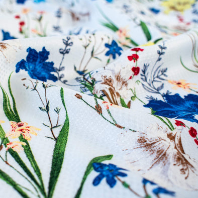 Italian crafted, soft white, viscose woven fabric by the yard. Gorgeous lilies and other florals in shades of peach, red, ivory and a vibrant blue with a fine jacquard pattern. Close up image.