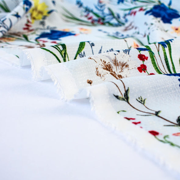 Italian crafted, soft white, viscose woven fabric by the yard. Gorgeous lilies and other florals in shades of peach, red, ivory and a vibrant blue with a fine jacquard pattern. Image of fabric selvedge.