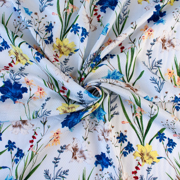 Italian crafted, soft white, viscose woven fabric by the yard. Gorgeous lilies and other florals in shades of peach, red, ivory and a vibrant blue with a fine jacquard pattern. Image of fabric body.