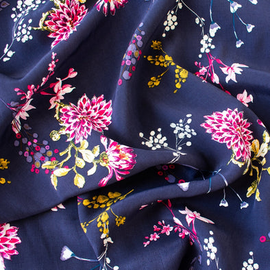 Italian Navy Floral Viscose Crepe fabric by the yard. This fabric features a beautiful design of vibrant blooms in shades of pink, and delicate sprays of flowers in soft white and ochre set against a navy background. The elegant drape and slightly textured crepe-like hand is perfect for your next high-end garment. Close up image.