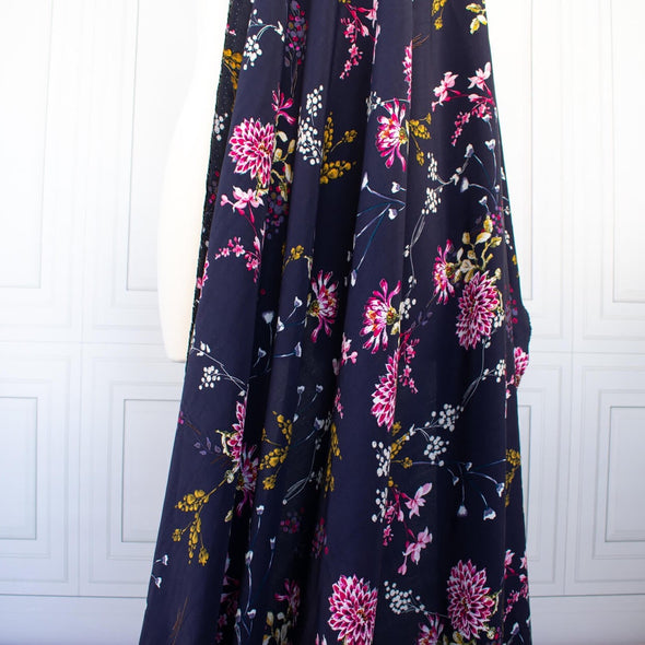 Italian Navy Floral Viscose Crepe fabric by the yard. This fabric features a beautiful design of vibrant blooms in shades of pink, and delicate sprays of flowers in soft white and ochre set against a navy background. The elegant drape and slightly textured crepe-like hand is perfect for your next high-end garment. Image of fabric drape.