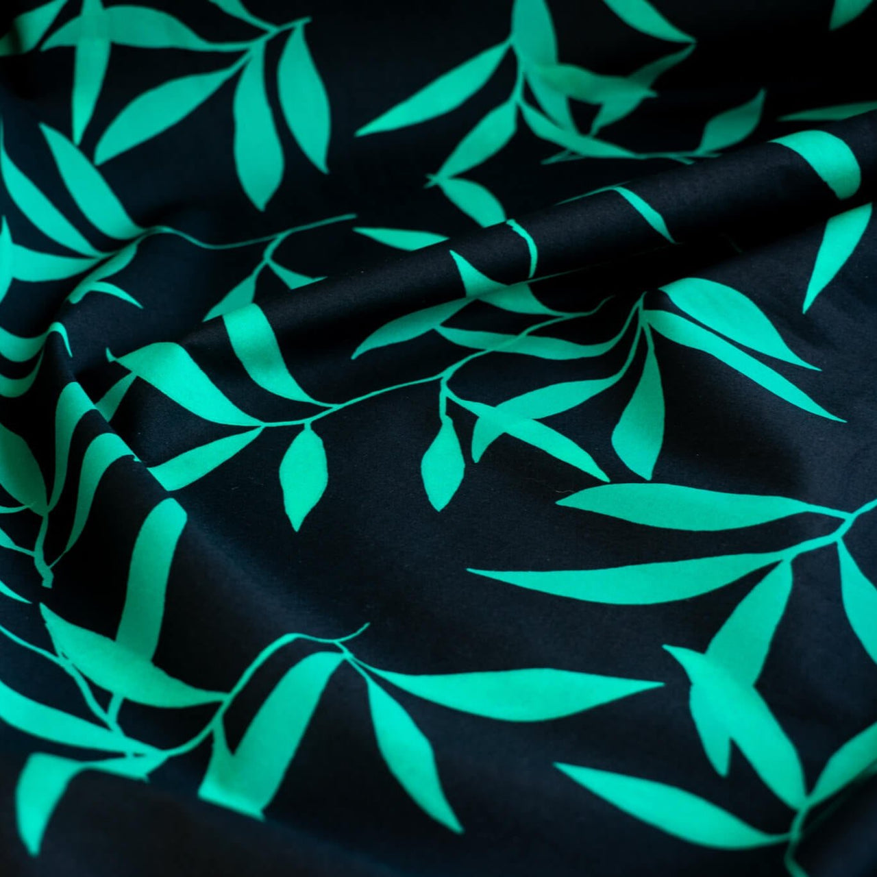 Carolina' cotton poplin designer fabric by the yard. This Italian poplin is light and billowy, perfect for sewing up tops, dresses and skirts. Made with stretch for ultimate comfort, this fabric is perfect if you want to add a touch of the exotic with its turquoise bamboo leaf print set against a black background. Close up image.