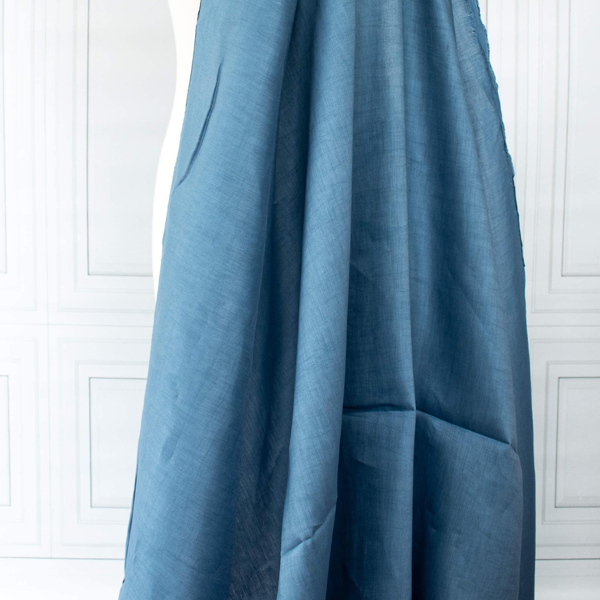 Image of Italian linen and viscose blend woven fabric by the yard in a soft Aegean blue draped on dress form against a white paneled wall background.