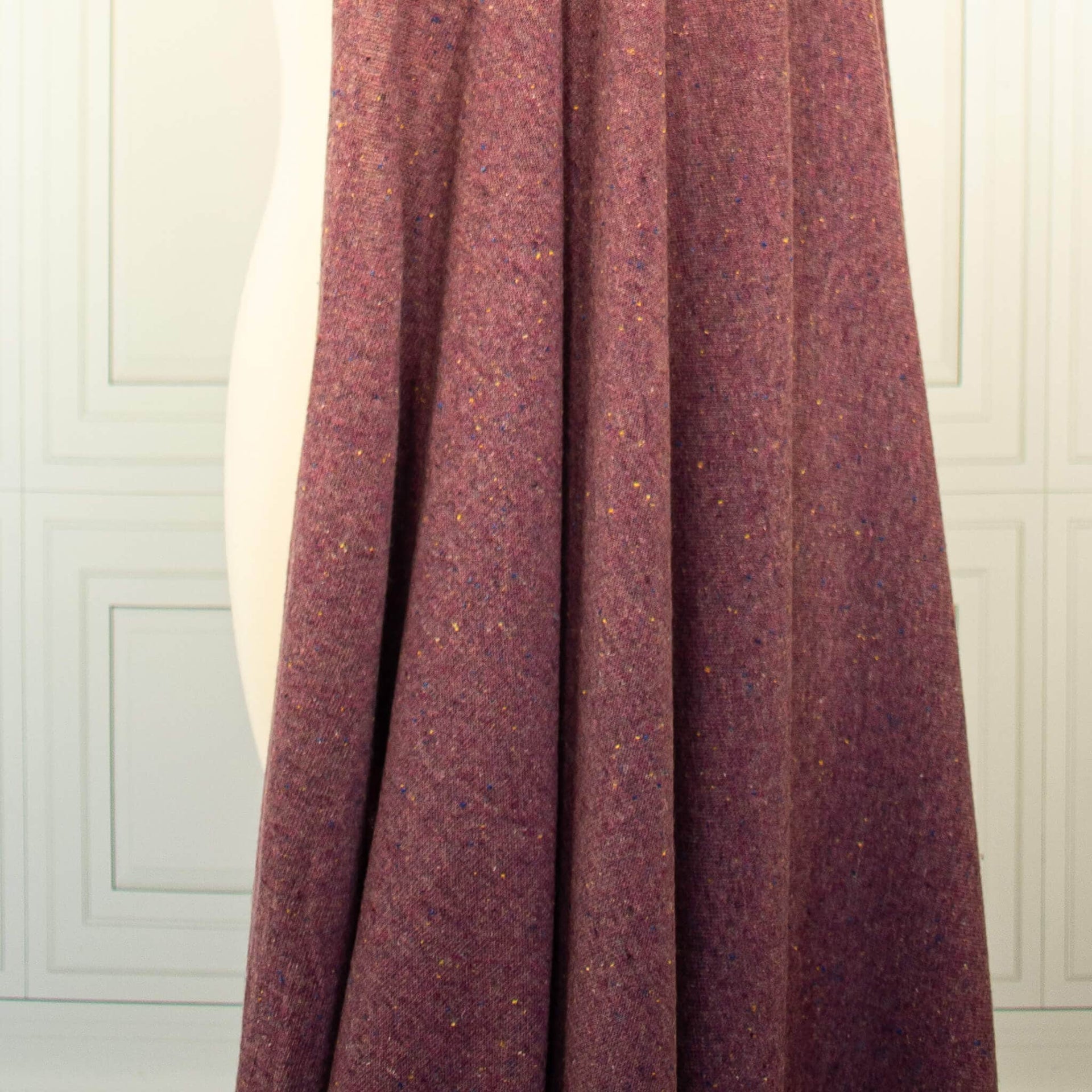 A close-up of Fine Italian Wool/Silk Blend Knit fdraped fabric in a heathered Violet Iris color with subtle multicolored flecks, shown against a white paneled wall background.