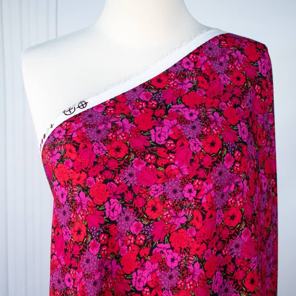 Italian woven viscose crepe fabric by the yard. Beautiful blooms are perfect when you want to add a burst vibrant color! Richly printed in shades of red, pink and lilac with vibrant green leaves set against a black background.  Image of fabric on dressform.