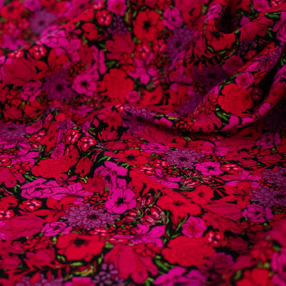 Italian woven viscose crepe fabric by the yard. Beautiful blooms are perfect when you want to add a burst vibrant color! Richly printed in shades of red, pink and lilac with vibrant green leaves set against a black background.  Close of image.