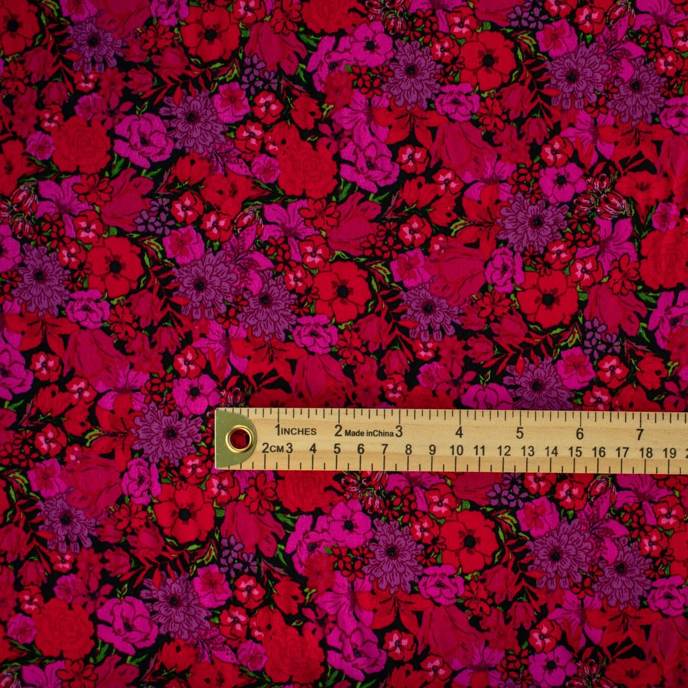 Italian woven viscose crepe fabric by the yard. Beautiful blooms are perfect when you want to add a burst vibrant color! Richly printed in shades of red, pink and lilac with vibrant green leaves set against a black background.  Image of fabric pattern scale with ruler.