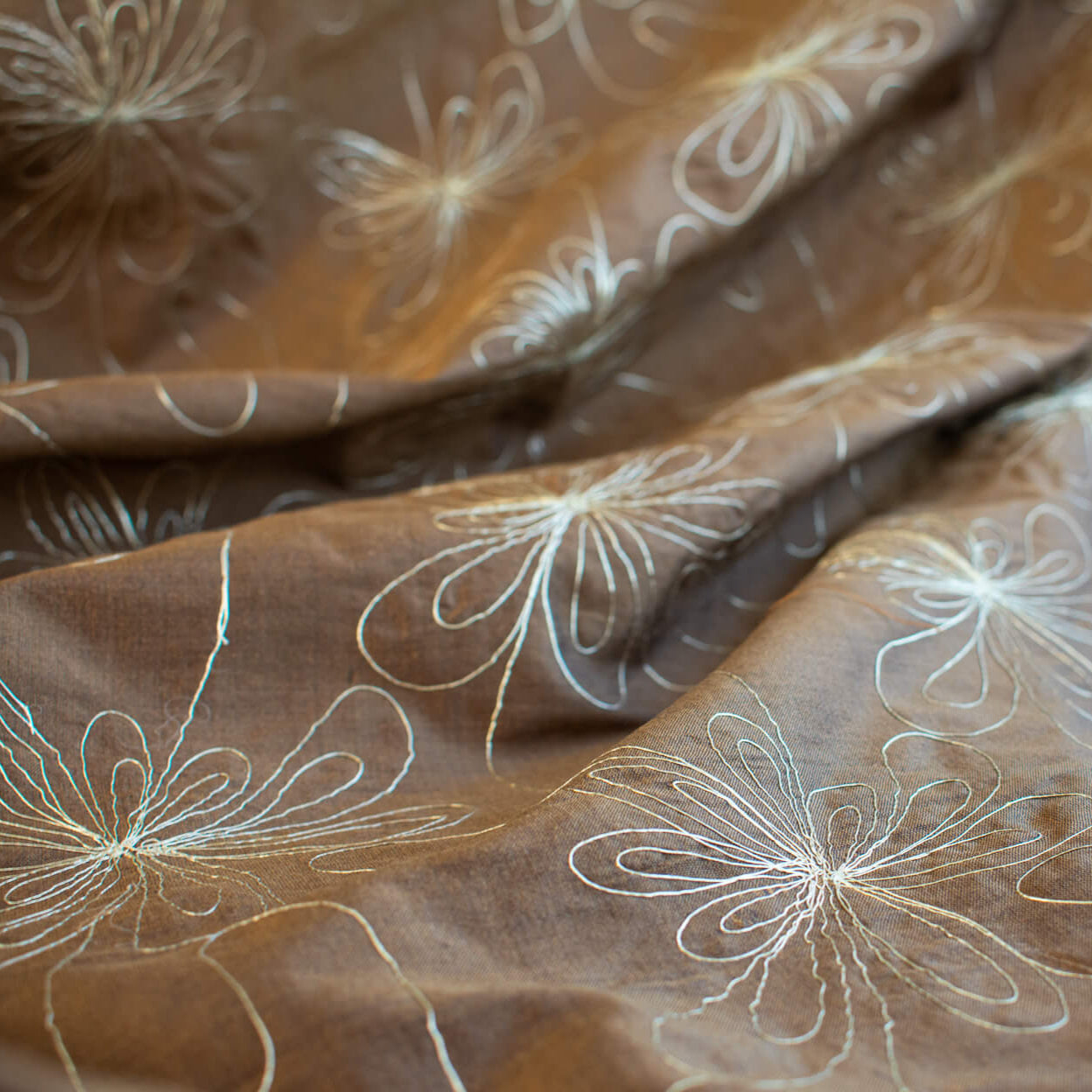 100% silk shantung fabric by the yard featuring a metallic embroidered floral design in a modern and playful hand-drawn style. In a rich hazelnut brown hue, this fabric offers a crisp and light textured hand, with a soft sheen for added elegance. Close up image.