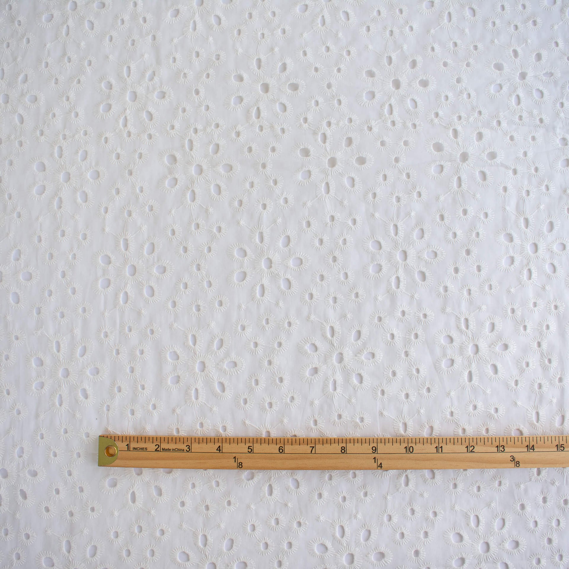 Image of  Italian Cotton San Gallo Lace in soft white, a delicate broderie anglaise fabric shown flat with a wooden ruler at the bottom for scale.