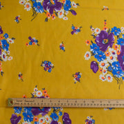 Image of Italian Cotton Floral Voile fabric by the yard in saffron yellow featuring large flowers in purple, blue, orange, and white shown with a wooden ruler across the middle for scale.