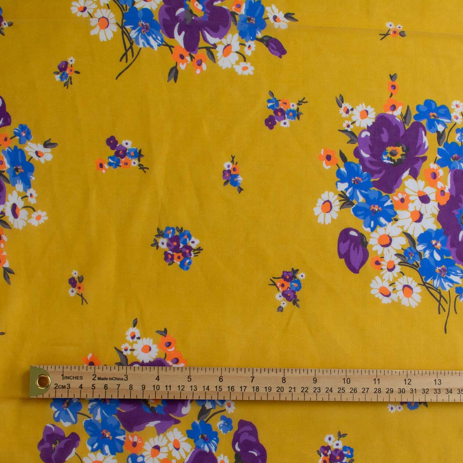 Image of Italian Cotton Floral Voile fabric by the yard in saffron yellow featuring large flowers in purple, blue, orange, and white shown with a wooden ruler across the middle for scale.