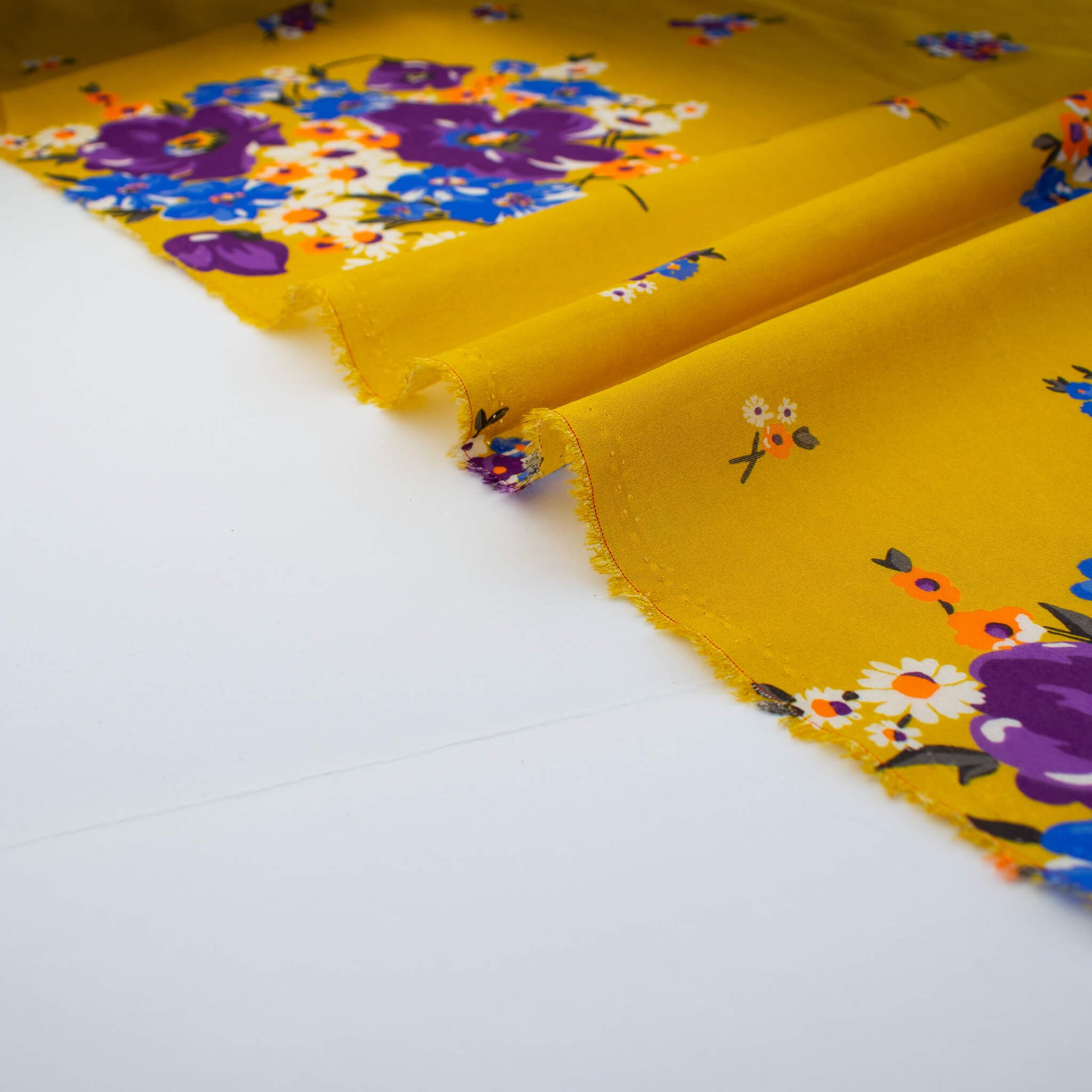 A close-up of Italian Cotton Floral Voile fabric selvedge in saffron yellow featuring large flowers in purple, blue, orange, and white.