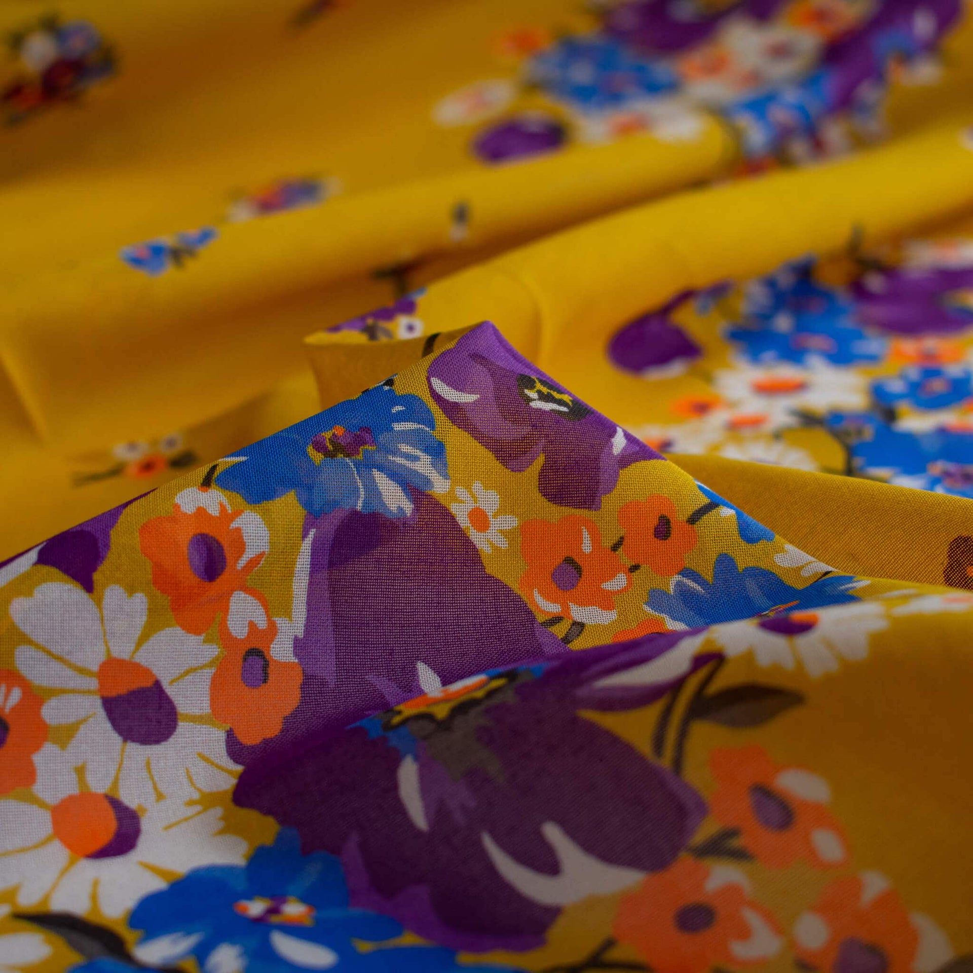 A close-up of draped Italian Cotton Floral Voile fabric by the yard in saffron yellow featuring large flowers in purple, blue, orange, and white.