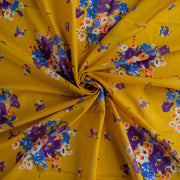 A close-up of Italian Cotton Floral Voile fabric by the yard in saffron yellow featuring large flowers in purple, blue, orange, and white arranged in a spiral to demonstrate fabric fullness