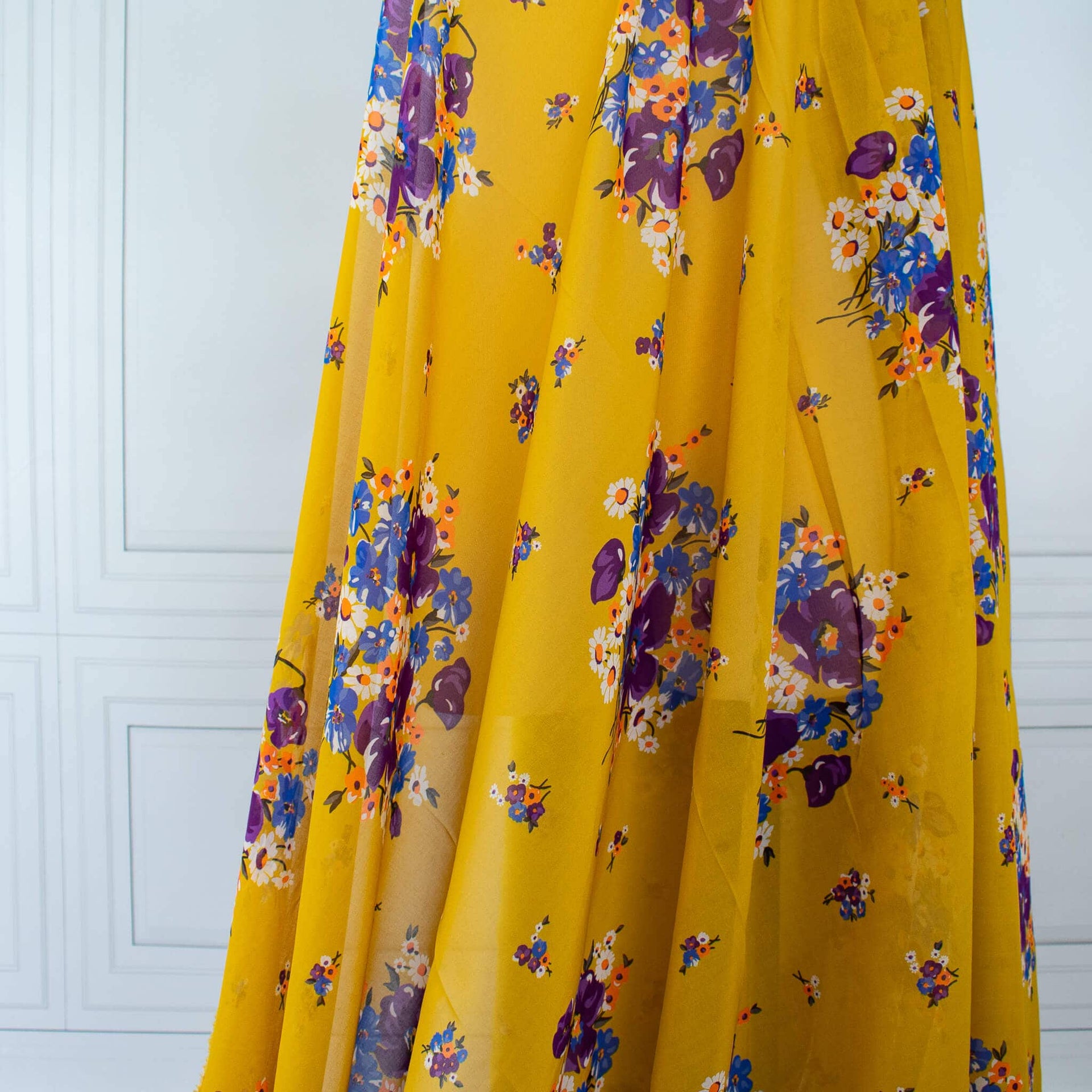 A close-up of draped Italian Cotton Floral Voile fabric by the yard in saffron yellow featuring large flowers in purple, blue, orange, and white.
