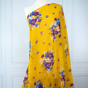 A close-up of draped Italian Cotton Floral Voile fabric by the yard in saffron yellow featuring large flowers in purple, blue, orange, and white draped  over a dress form 