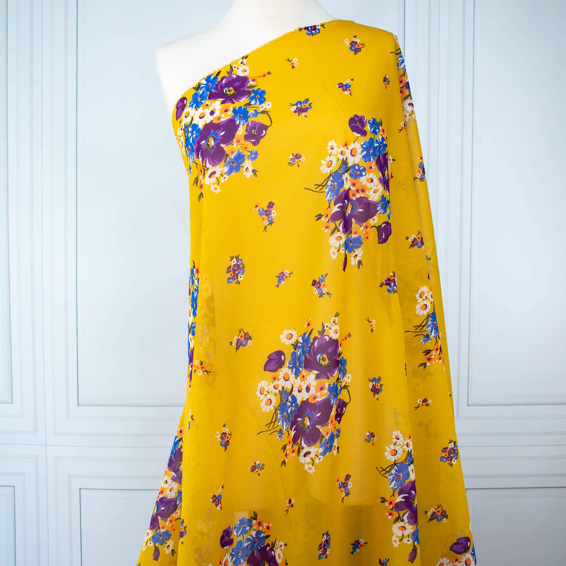A close-up of draped Italian Cotton Floral Voile fabric by the yard in saffron yellow featuring large flowers in purple, blue, orange, and white draped  over a dress form 