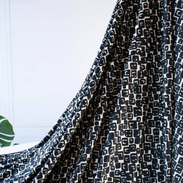 Designer Italian Matte Jersey Knit Fabric in a modern geometric print of abstract black squares se against a dark creme background - picture shows fabric drape.