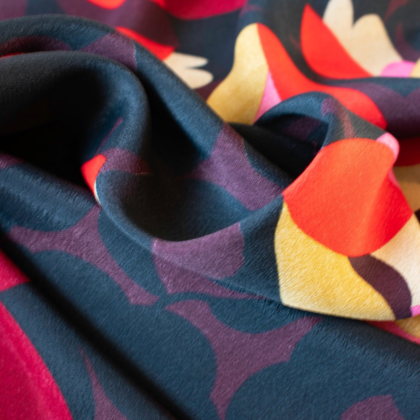 Tulip petals in red, ochre, pink and maroon float above off black daisies in a rich background of aubergine. Floral viscose crepe from Italy has a lovely drape. Close up photo.