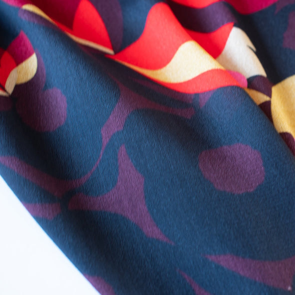 Tulip petals in red, ochre, pink and maroon float above off black daisies in a rich background of aubergine. Floral viscose crepe from Italy has a lovely drape. Close up photo.