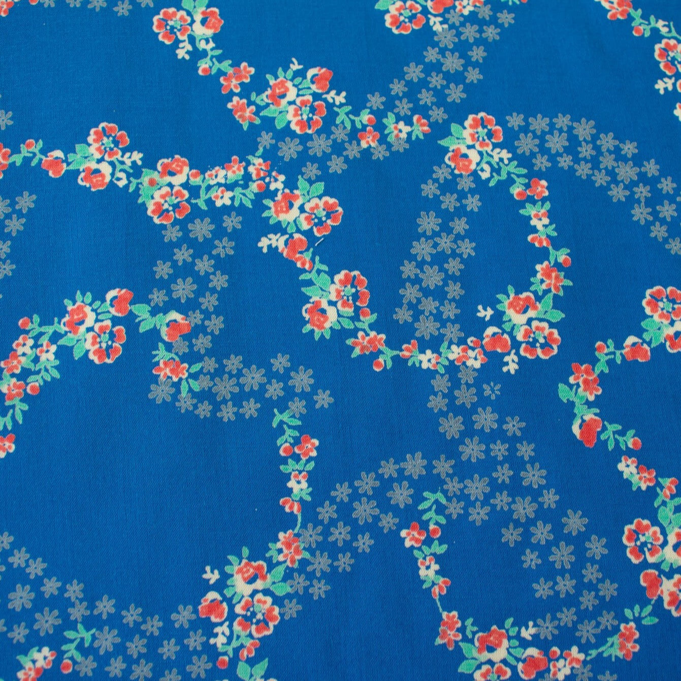 A stunning blue rayon floral challis will have you dreaming of summer strolls, taking in the sunshine and feeling so fine.  Soft and airy, perfect for lazy day dresses, billowy tops, or a simple cami and skirt.    Trailing light grey daises and red flowers with light turquoise leaves trail a delicate path against a medium blue background.   Image of fabric print design.