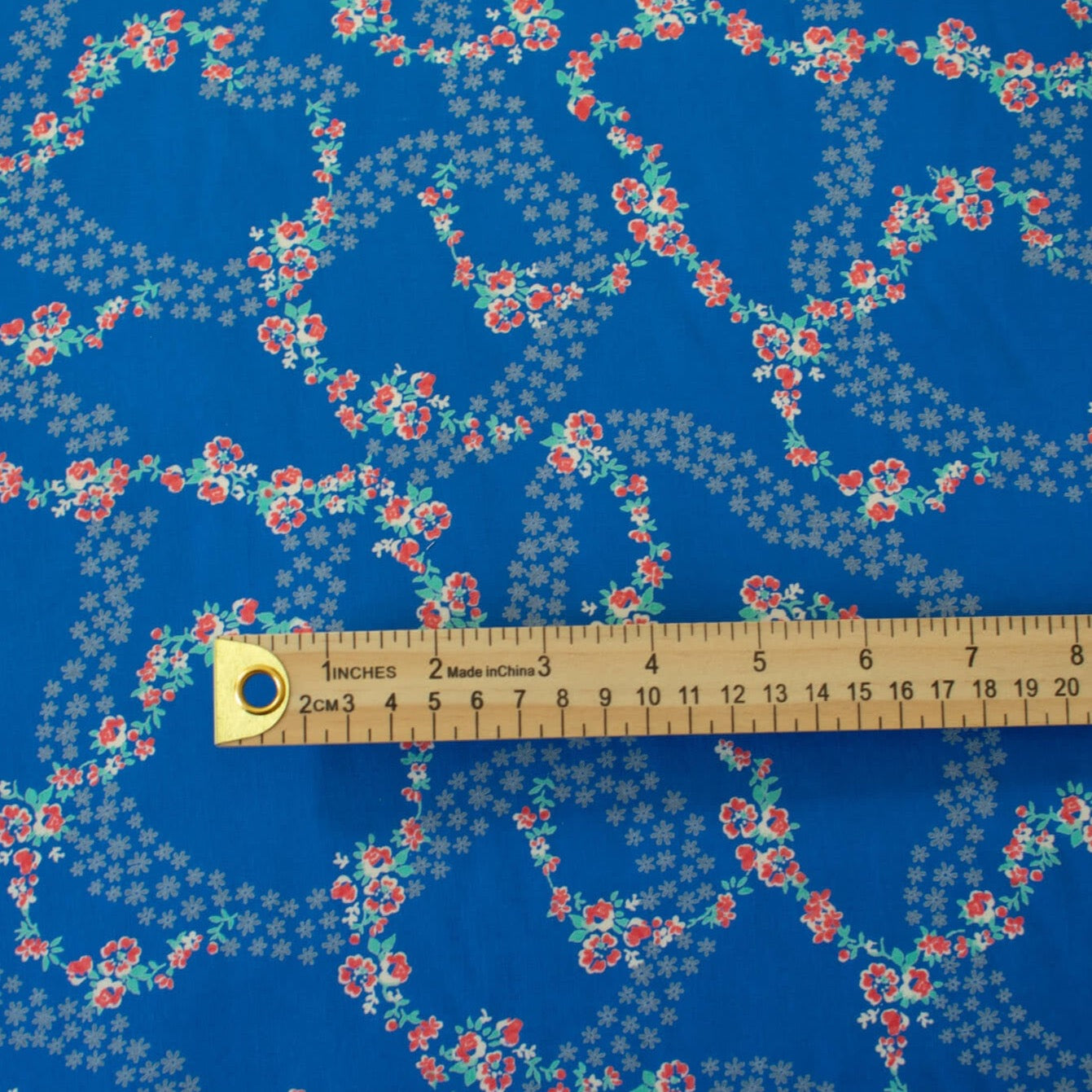 A stunning blue rayon floral challis will have you dreaming of summer strolls, taking in the sunshine and feeling so fine.  Soft and airy, perfect for lazy day dresses, billowy tops, or a simple cami and skirt.    Trailing light grey daises and red flowers with light turquoise leaves trail a delicate path against a medium blue background.   Image of fabric with ruler for design scale.