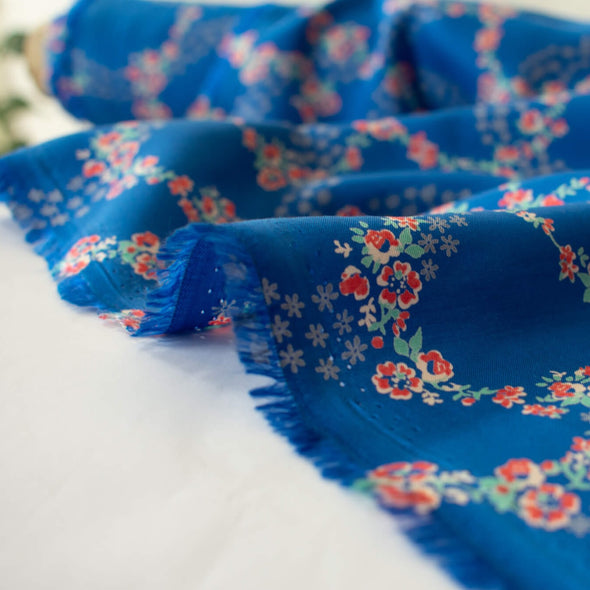 A stunning blue rayon floral challis will have you dreaming of summer strolls, taking in the sunshine and feeling so fine.  Soft and airy, perfect for lazy day dresses, billowy tops, or a simple cami and skirt.    Trailing light grey daises and red flowers with light turquoise leaves trail a delicate path against a medium blue background.   Image of selvedge.