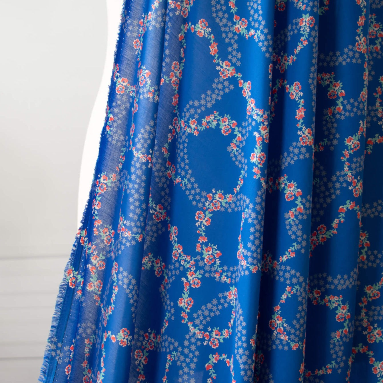 A stunning blue rayon floral challis will have you dreaming of summer strolls, taking in the sunshine and feeling so fine.  Soft and airy, perfect for lazy day dresses, billowy tops, or a simple cami and skirt.    Trailing light grey daises and red flowers with light turquoise leaves trail a delicate path against a medium blue background.   Image of fabric drape.